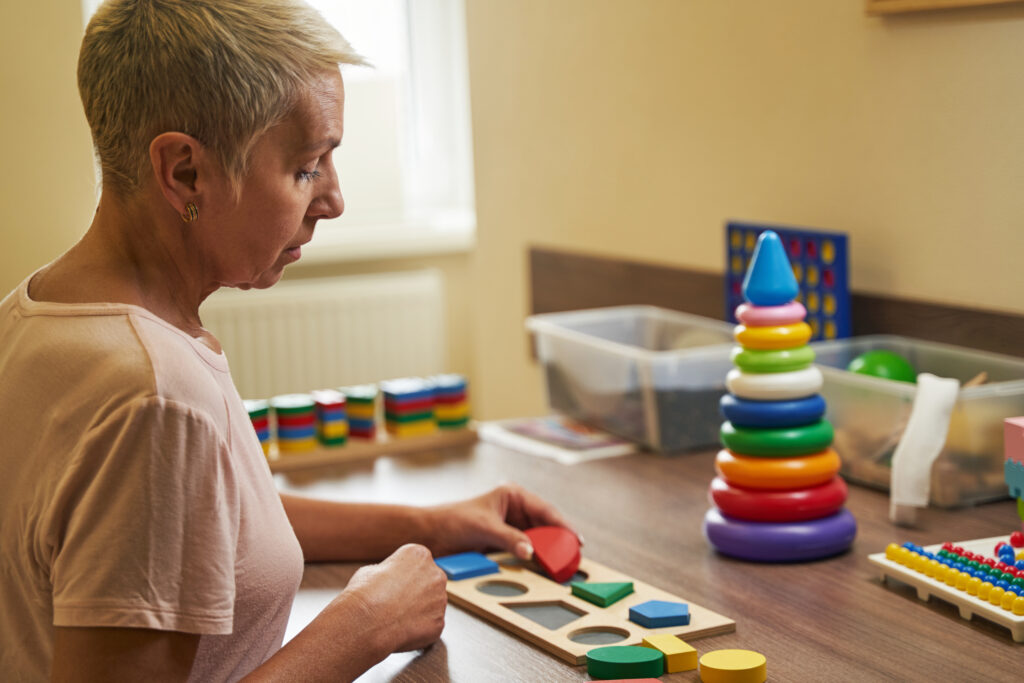 Photo of middle-aged woman on physical therapy with wooden puzzles after suffering from stroke in neurological rehabilitation facility