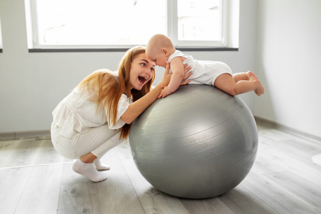 Happy mother with baby girl in exercise ball at home in living room. Occupational therapy.